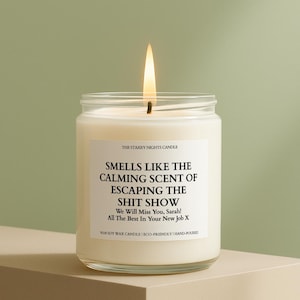May include: A lit, clear glass jar candle with a white label. The label reads "Smells Like The Calming Scent of Escaping The Shit Show." The candle is on a beige surface against a green background. The candle is hand-poured.