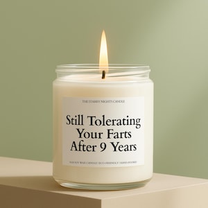 May include: A lit, white candle in a clear glass jar. The label reads "Still Tolerating Your Farts After 9 Years." The candle is on a neutral-toned surface against a green background. The text "THE STARRY NIGHTS CANDLE" is above the main text.