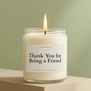 May include: A lit soy wax candle in a clear glass jar. The candle has a white label with the text "Thank You for Being a Friend". The candle is on a neutral-toned surface against a green background.