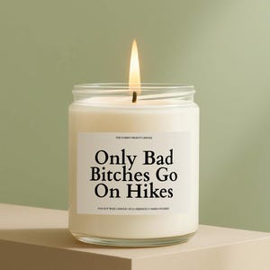 May include: A lit, white candle in a clear glass jar. The label reads "Only Bad Bitches Go On Hikes." The candle is on a neutral-toned surface against a green background. The text "THE STARRY NIGHTS CANDLE" is above the text on the label.