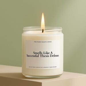 May include: A lit, white candle in a clear glass jar. The label reads "Smells Like A Successful Thesis Defense." The candle is hand-poured and made with soy wax. The background is a light green.