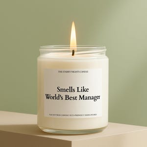 May include: A lit, white candle in a clear glass jar. The label reads "Smells Like World's Best Manager." The candle is labeled "The Starry Nights Candle" and is made with soy wax. The flame is bright.