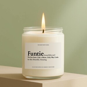 May include: A lit, white soy wax candle in a clear glass jar. The label reads "Funtie" with a definition. The candle is hand-poured and eco-friendly. The flame is bright against a green background.