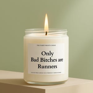 May include: A burning candle in a clear glass jar. The label says "Only Bad Bitches are Runners", with "The Starry Nights Candle" above and "100% Soy Wax Candle | Eco-Friendly | Hand-poured" below. The candle is on a beige surface.