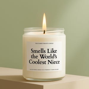May include: A lit, white soy wax candle in a clear glass jar. The label reads "Smells Like the World's Coolest Niece." The candle is hand-poured and eco-friendly. The background is a soft green.