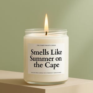 May include: A lit candle in a clear glass jar. The candle has a white label with the text "Smells Like Summer on the Cape". The candle is on a beige surface against a green background. The text "THE STARRY NIGHTS CANDLE" is above the main text.