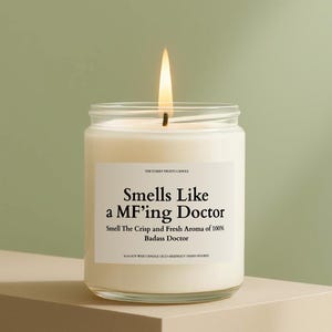 Puede incluir: Una vela encendida de cera de soja blanca en un tarro de cristal transparente. La etiqueta dice "Smells Like a MF'ing Doctor". La vela es vertida a mano y ecológica, con un aroma fresco.
