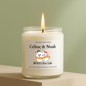 May include: A lit, white soy wax candle in a glass jar. The label reads "Celine & Noah" above a cartoon couple and "BOO's For Life." The candle is hand-poured and eco-friendly.