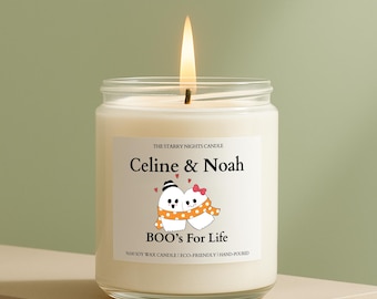 May include: A lit, white soy wax candle in a glass jar. The label reads "Celine & Noah" above a cartoon couple and "BOO's For Life." The candle is hand-poured and eco-friendly.
