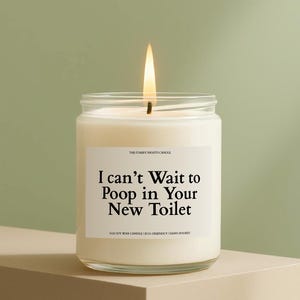 May include: A lit candle in a clear glass jar with a white label. The label reads "I can't Wait to Poop in Your New Toilet". The candle is on a neutral-colored surface against a green background.
