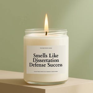 May include: A lit, white soy wax candle in a clear glass jar. The label reads "Smells Like Dissertation Defense Success." The candle is hand-poured and eco-friendly. The background is a soft green.