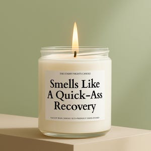 May include: A lit candle in a clear glass jar with a white label. The label reads "Smells Like A Quick-Ass Recovery" in black text. The candle is on a beige surface against a green background. The text "THE STARRY NIGHTS CANDLE" is above the main text.