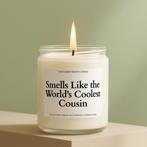 May include: A lit candle in a clear glass jar with a white label that reads "Smells Like the World's Coolest Cousin." The candle is labeled "The Starry Nights Candle" and is made of soy wax.