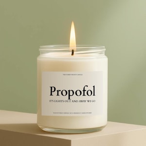 May include: A lit, white soy wax candle in a clear glass jar. The label reads "Propofol, It's Lights Out and Away We Go." The candle is hand-poured and eco-friendly.