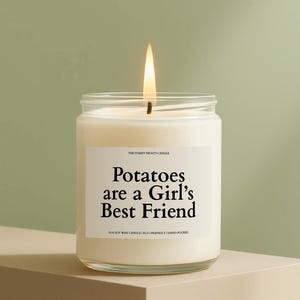 May include: A lit soy wax candle in a clear glass jar. The candle has a white label with the text "Potatoes are a Girl's Best Friend." The candle is hand-poured and eco-friendly. The background is a light green.
