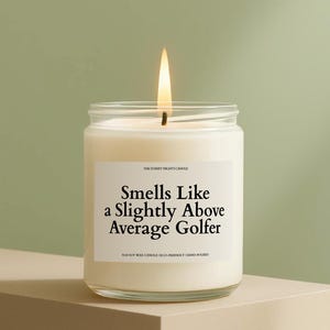 Peut inclure: Une bougie en cire de soja blanche allumée dans un pot en verre transparent. L'étiquette indique "Smells Like a Slightly Above Average Golfer". La bougie est coulée à la main et écologique.