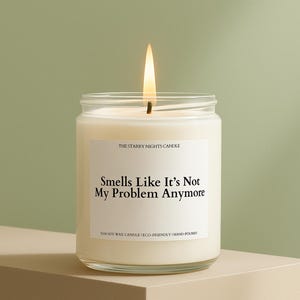 May include: A lit, white candle in a clear glass jar. The label reads "Smells Like It's Not My Problem Anymore" with "The Starry Nights Candle" above and "%100 Soy Wax Candle | Eco-Friendly | Hand-poured" below. The candle sits on a beige surface.