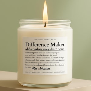 May include: A clear glass jar candle with a white wax fill and a lit wick. The label reads: THE STARRY NIGHTS CANDLE, Difference Maker, /dif-er-uhns mey-ker/ noun, a dedicated person who can make a big impact, See also: Mrs Johnson, %100 SOY WAX.