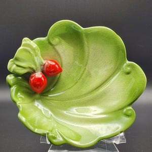 May include: A ceramic, leaf-shaped dish in shades of green, with two glossy red berry accents. The dish has a textured surface and is displayed on a clear stand against a dark background. The design is reminiscent of nature.