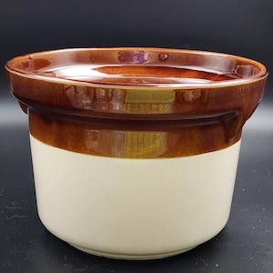 May include: A ceramic crock with a two-tone design. The top and rim are a glossy brown, with a wide band of brown separating the top from the off-white base. The crock has a rounded shape and a smooth, reflective surface.