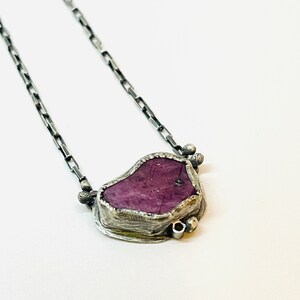 May include: A silver chain necklace with a purple gemstone pendant. The pendant is set in a silver frame with small silver beads around the edge.