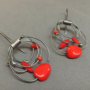 May include: A pair of silver-toned wire earrings. Each earring has a series of looped wires, with bright red, irregularly shaped beads. The earrings have a hook closure. The design is abstract and modern.