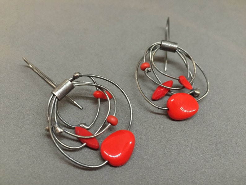 May include: A pair of silver-toned wire earrings. Each earring has a series of looped wires, with bright red, irregularly shaped beads. The earrings have a hook closure. The design is abstract and modern.