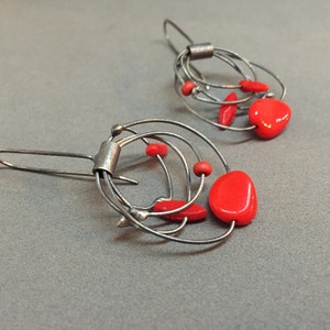 May include: A pair of silver wire earrings featuring vibrant red beads. The earrings have an abstract design with multiple wire loops and small red accents. The design creates a modern, artistic look, suitable for everyday wear.