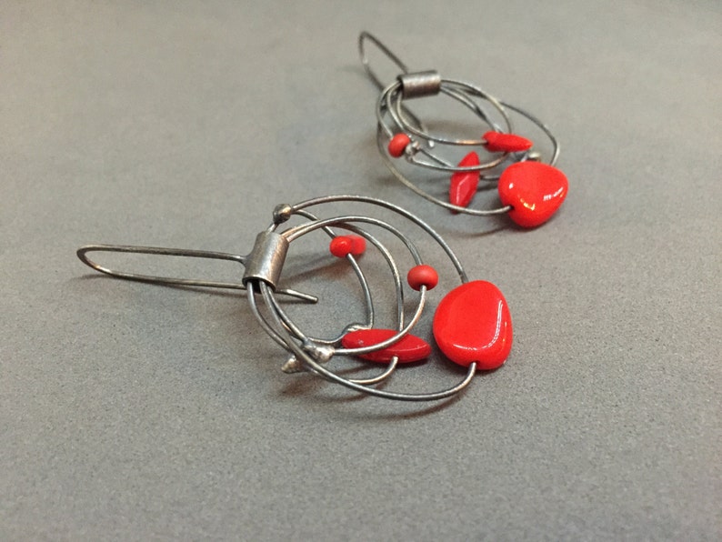 May include: A pair of silver wire earrings featuring vibrant red beads. The earrings have an abstract design with multiple wire loops and small red accents. The design creates a modern, artistic look, suitable for everyday wear.