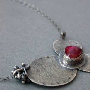 May include: A silver-toned necklace featuring a pendant with a large, oval, ruby-red gemstone set in a textured metal setting. The pendant is attached to a delicate chain. The design includes three textured, circular elements.