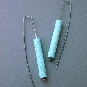 May include: A pair of handmade earrings featuring long, silver-tone ear wires. Each earring has a cylindrical design made of stacked, light blue paper discs, with a small silver bead at the bottom. The earrings are displayed against a gray background.