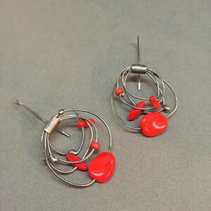 May include: Abstract earrings with a silver wire design, incorporating multiple loops and red, oval-shaped details. The earrings have a post closure and a contemporary, artistic style. The red accents add a pop of colour.