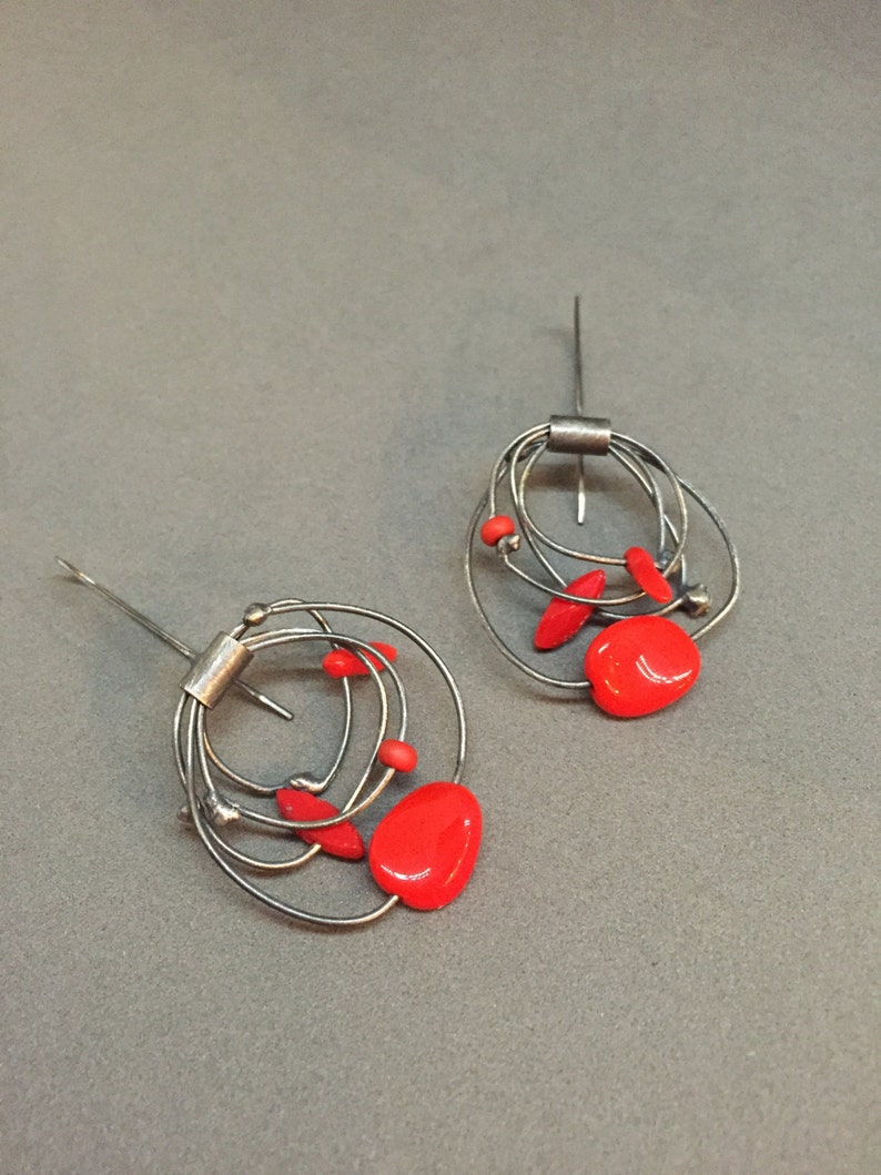 May include: Abstract earrings with a silver wire design, incorporating multiple loops and red, oval-shaped details. The earrings have a post closure and a contemporary, artistic style. The red accents add a pop of colour.