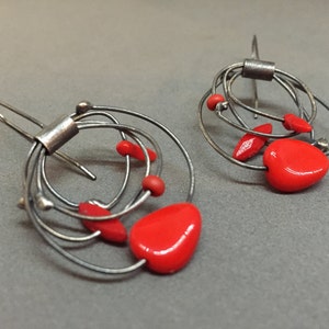 May include: A pair of abstract silver earrings with red heart-shaped and oval accents. The earrings have a wire design with multiple loops and a hook closure. The red elements provide a striking contrast to the silver metal.