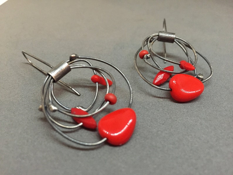 May include: A pair of abstract silver earrings with red heart-shaped and oval accents. The earrings have a wire design with multiple loops and a hook closure. The red elements provide a striking contrast to the silver metal.