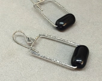 May include: A pair of silver-toned rectangular earrings with a hammered texture. Each earring features a glossy, black, oblong bead suspended within the frame. The earrings have a simple hook closure.