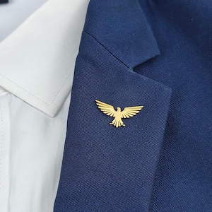 May include: A gold-colored lapel pin in the shape of a hawk or eagle with its wings spread. The pin is on a dark blue blazer.