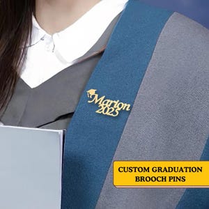 Puede incluir: Un broche de graduación personalizado de color dorado con el nombre "Marion" y el año "2025". El broche está sujeto a una banda de graduación azul y gris. El texto "CUSTOM GRADUATION BROOCH PINS" está en un rectángulo amarillo.