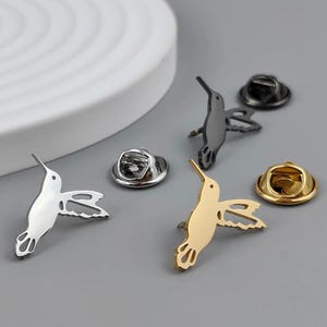 May include: Four hummingbird pins with different colored finishes. The pins are silver, gold, black, and black with a silver finish. Each pin has a safety clasp on the back.