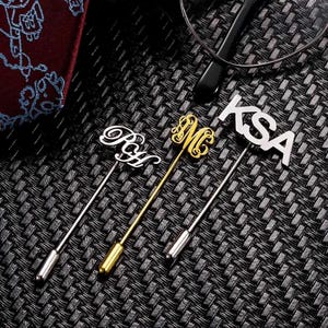 Może przedstawiać: Trzy srebrne spinki do krawata z różnymi wygrawerowanymi inicjałami. Inicjały to "KSA", "M" i "RYL".