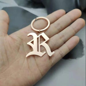 May include: Rose gold keychain with a large, ornate letter R charm. The charm is in a gothic font style.