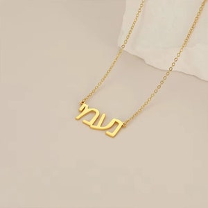 May include: A gold-colored necklace with a chain and a pendant featuring Hebrew script. The pendant is in a sans-serif font and is centered on the chain. The necklace is displayed on a light-colored surface.