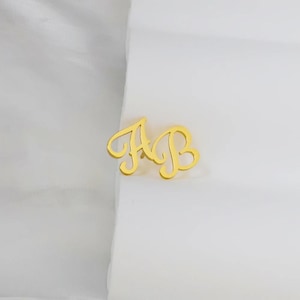 Pu&ograve; includere: Spilla con monogramma dorata con le lettere intrecciate "A" e "B" in corsivo. La spilla &egrave; presentata su uno sfondo bianco, evidenziando il suo design elegante. Questo accessorio &egrave; adatto per personalizzare abiti o accessori.