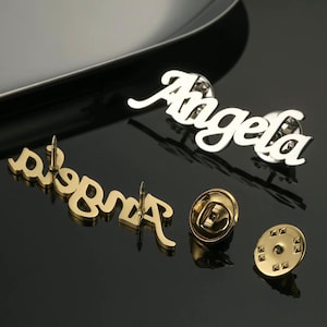 May include: Two gold-tone metal name pins with the name "Angela" spelled out in cursive. The pins have a safety pin closure.