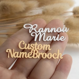 May include: Two custom name brooches. One is silver and reads "Rannou Marie" in cursive. The other is gold and says "Custom Name Brooch". Both are displayed on a hand.