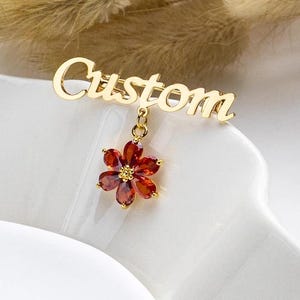 May include: Gold-tone brooch featuring the word "Custom" and a red flower charm.