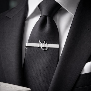 Può includere: Primo piano di una cravatta nera con fermacravatta argentato. Il fermacravatta presenta le lettere "N" e "G" in un design stilizzato. La cravatta fa parte di un abito nero con una camicia bianca.