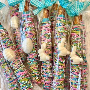 Op de afbeelding: Pretzelstaven bedekt met kleurrijke snoepcoating, versierd met pastelkleurige hagelslag en witte chocolade paaseieren en konijnenvormen. Elke pretzel is verpakt in doorzichtig cellofaan en vastgebonden met een blauw-wit geruit lint.