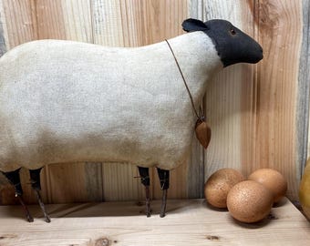 May include: A handcrafted sheep figurine with a black head and legs, and a cream-colored body. The sheep has a brown pendant and is next to three brown eggs. A yellow knitted object and a wooden chicken figurine are also present.
