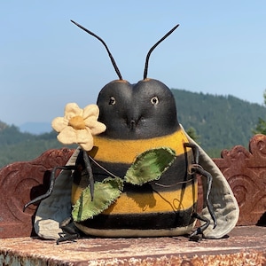 May include: A fabric bee figurine featuring a rounded body with horizontal black and yellow stripes, two thin antennae, and light-colored wings. The bee holds a small, light-colored fabric flower in one hand and has a green fabric leaf attached.